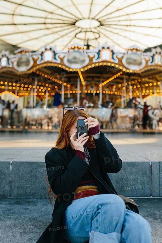 A woman with long hair uses a vintage camera to capture the lively atmosphere in Brooklyn Bridge Park. The colorful carousel and its lights create a festive backdrop