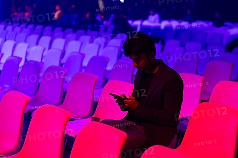 A indian man sits alone in an auditorium filled at a conference of AI in Amsterdam with colorful lights, focused on his smartphone. Empty chairs surround him, creating a contemplative scene