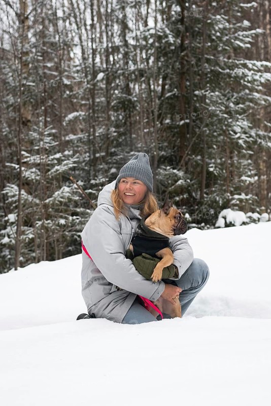 Woman in a winter coat and beanie happily embracing a dog in a snowy forest setting. The serene snow-covered landscape enhances the scene's tranquility
