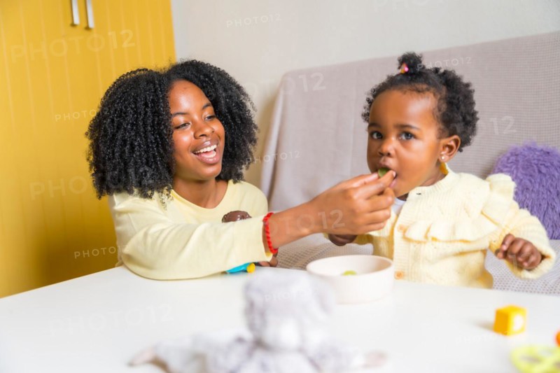 African mother feeding with hand her daughter in the living room at home