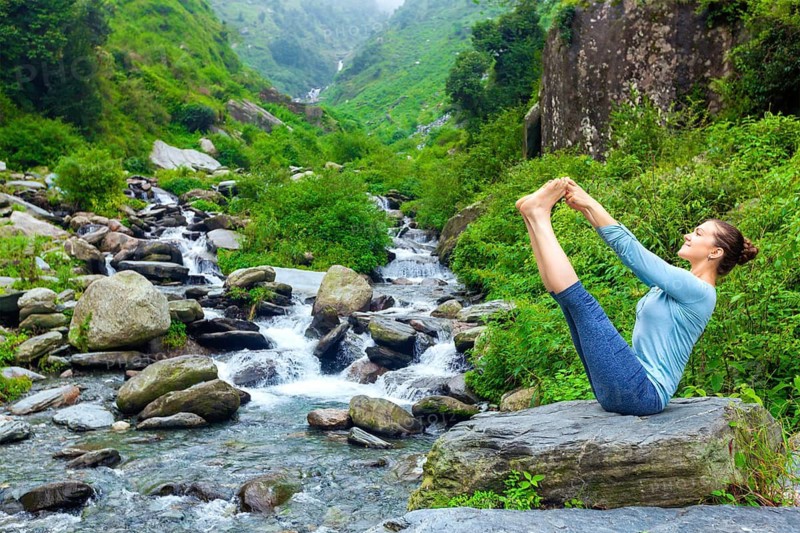 Yoga exercise outdoors, panorama of woman doing Ashtanga Vinyasa Yoga balance asana Ubhaya padangusthasana Big Double Toe Yoga Pose at tropical waterfall in Himalayas in India. Panoramic image