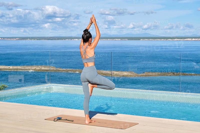 A woman stands in a yoga pose on a rooftop overlooking a serene ocean. Dressed in blue active wear, she enjoys tranquility and focus in a peaceful outdoor setting