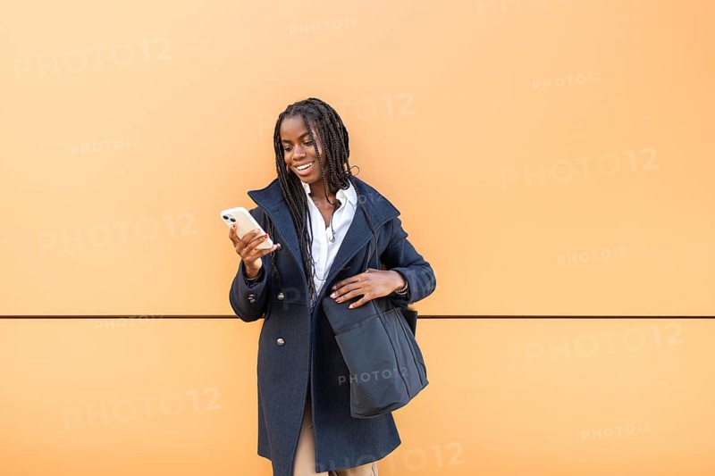 A young cheerful African American businesswoman with braids checks her smartphone, dressed in professional attire with a vibrant orange background