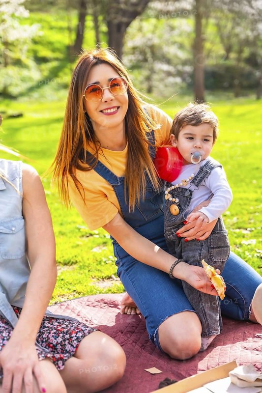 Young mother holding and feeding a slice of pizza to her baby while enjoying a picnic in a park with friends