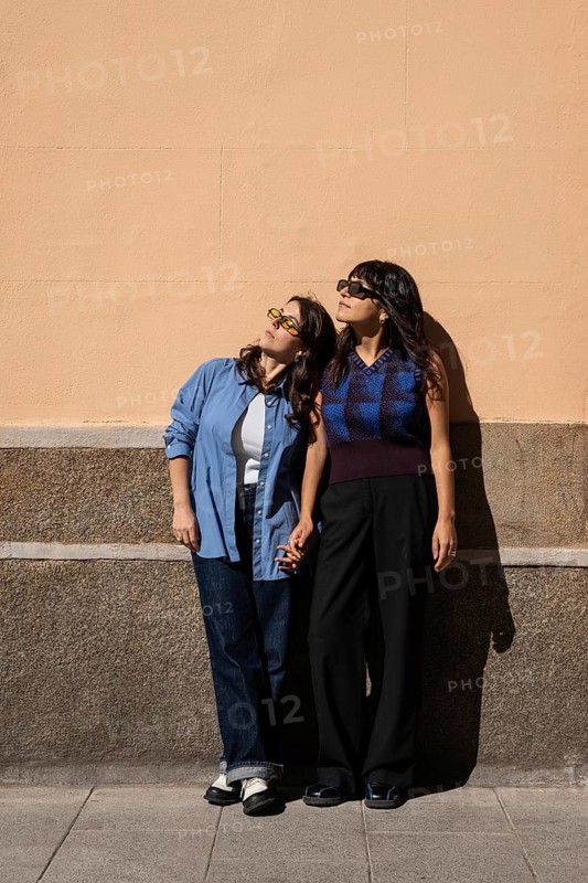 A lesbian couple stands outdoors, holding hands and basking in the sun. They celebrate love and diversity, reflecting a multiethnic connection with pride and joy