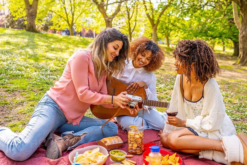 Three women relaxing on blanket, sharing food, playing guitar, and taking photos during sunny picnic in park