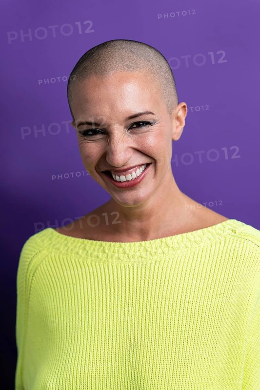 A middle-aged woman with a shaved head smiles warmly, wearing a neon green sweater against a vibrant purple background, conveying confidence and positivity