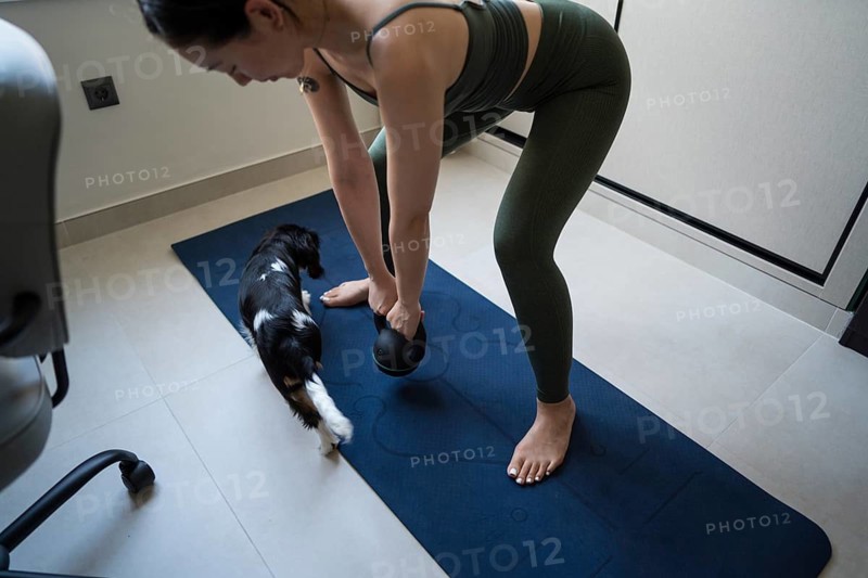 A young Asian woman attempts a home exercise routine on a yoga mat, but her playful dog interrupts, bringing a sense of joy to her workout space