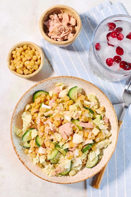 A fresh salad featuring tuna, corn, and cucumber on a striped cloth. Complemented by a drink with ice and berries, creating an appetizing and healthy meal setting