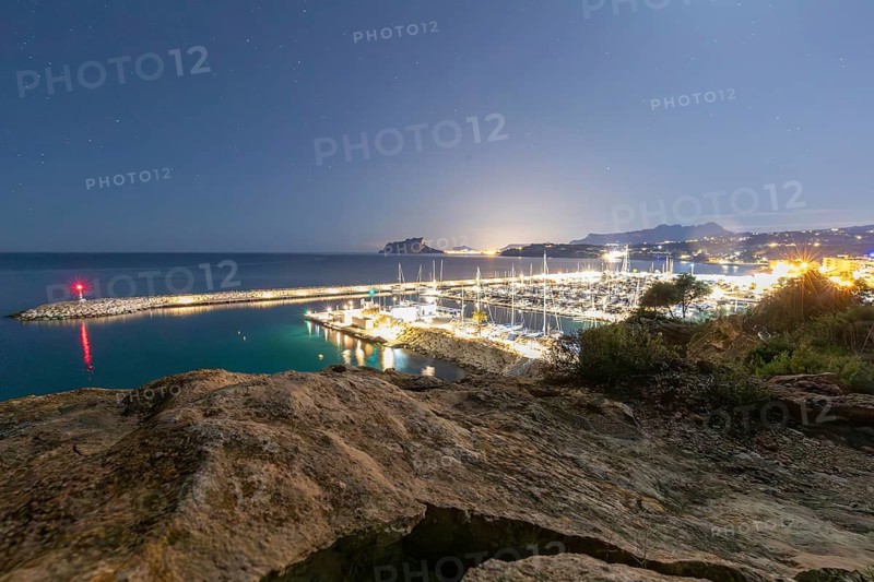Night descends on Costa Blanca, illuminating the harbor under a starlit sky. The Mediterranean Sea glimmers as the lights create a serene atmosphere, capturing natural beauty
