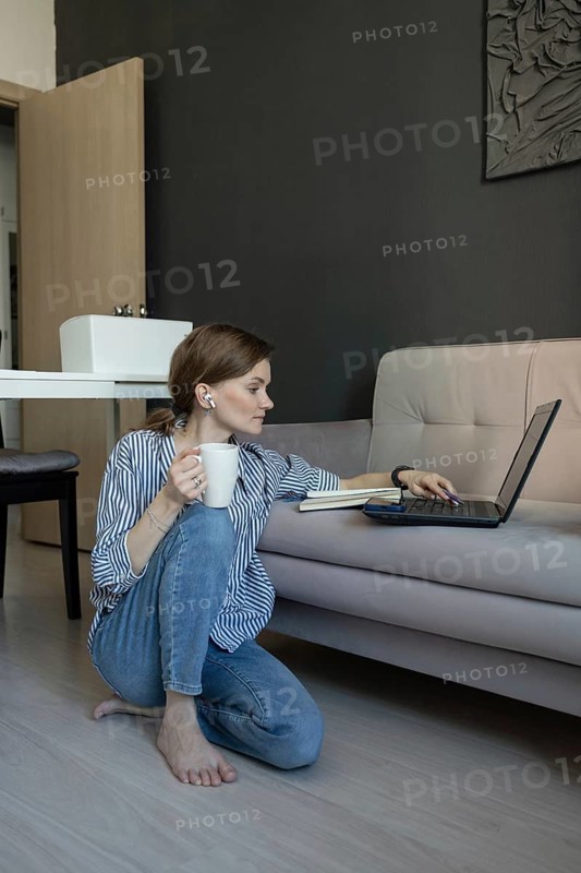 A woman sitting on the floor by a sofa, working on a laptop. She holds a coffee mug and listens with earbuds. The atmosphere is cozy and relaxed at home