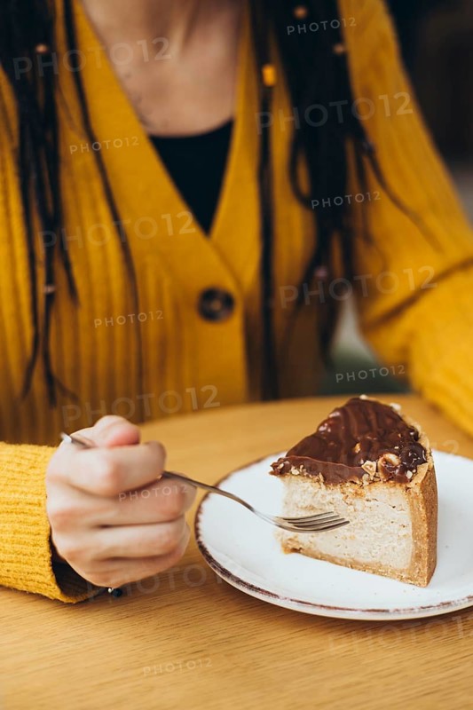 A person wearing a yellow sweater holds a fork, ready to enjoy a delicious slice of cake on a white plate The cake is topped with chocolate and nuts, set on a wooden table