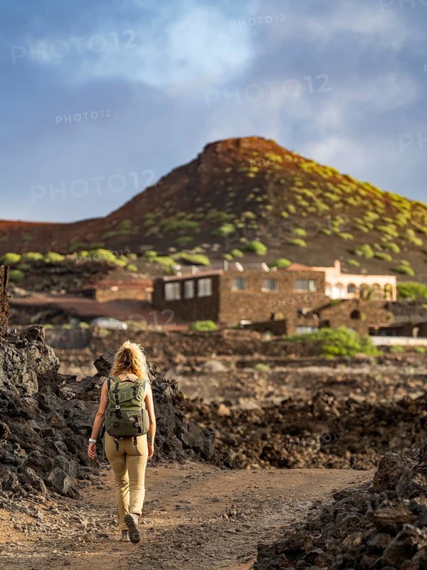 A woman with a backpack traverses the stark, volcanic terrain of Timanfaya National Park in Lanzarote, Canary Islands. The rugged landscape features vivid tints and a dormant volcano