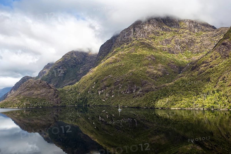 View from the deck of a cruise ship of an impressive mountain landscape, partly illuminated by sunlight. On the right, a narrow waterfall cascades down the rock face and is clearly reflected on the calm water surface. There is a single sailing ship on the shore, Chile, South America