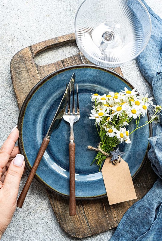 Top view of cropped unrecognizable hands of a woman adjust cutlery on a blue plate with summer daisies. Set on a rustic wooden board, creating an aesthetic interior decor