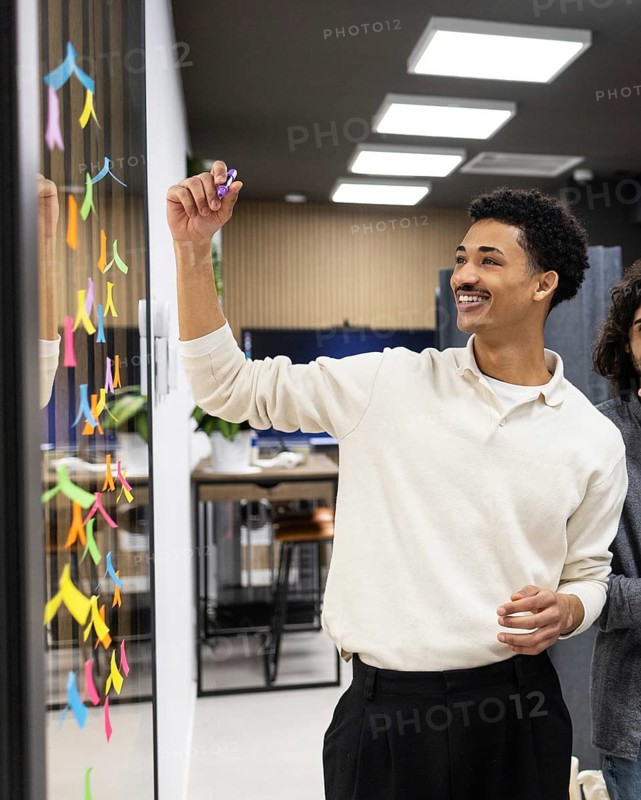 A professional in a modern office setting adds colorful sticky notes to a glass wall during a brainstorming session. The image captures teamwork and creativity in a corporate environment
