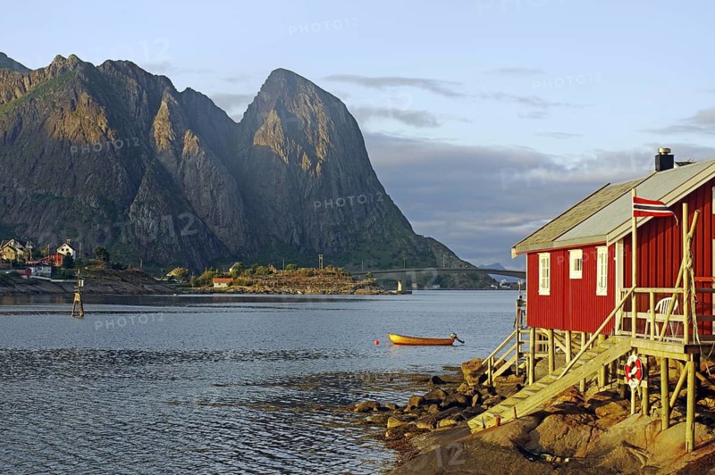 Red wooden hut with boat on the fjord shore, surrounded by mountains at sunset, Rorbuer, holiday accommodation, Reine, Moskenes, Lofoten, Nordland, Norway