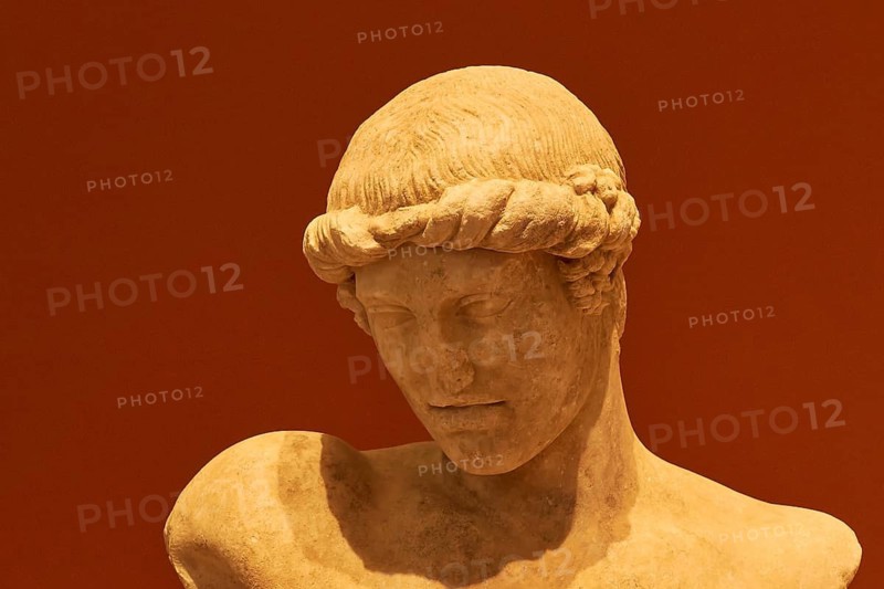 Ancient male head of a sculpture with detailed hairstyle against a warm background, possibly depicting the god Apollo, Patras Archaeological Museum, Patras, Peloponnese, Greece