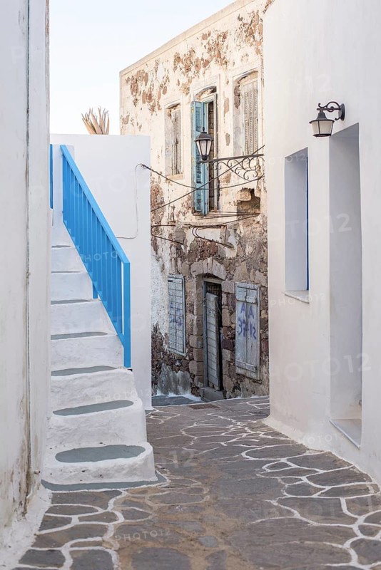 A charming narrow alleyway with rustic, weathered architecture in Plaka, Milos island, Greece. Features bright white painted stairs with a blue railing, leading to historic stone buildings