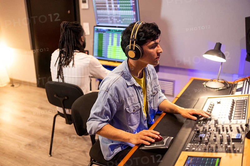 Two audio engineers work in a recording studio, wearing headphones and using mixing consoles and monitors to edit and produce music tracks. Focused, creative atmosphere