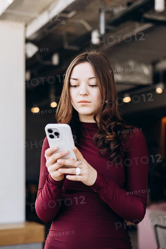A young female student with long hair smiles as she looks at her smartphone, wearing a maroon dress. She is indoors and appears to be enjoying herself while using her device