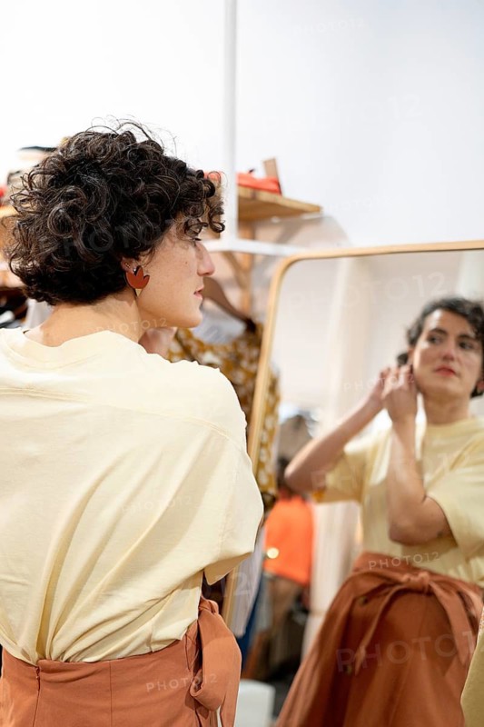 A woman stands in front of a large mirror, adjusting her earrings and admiring her reflection. The scene captures a moment of self care and personal expression in a cozy clothes shop