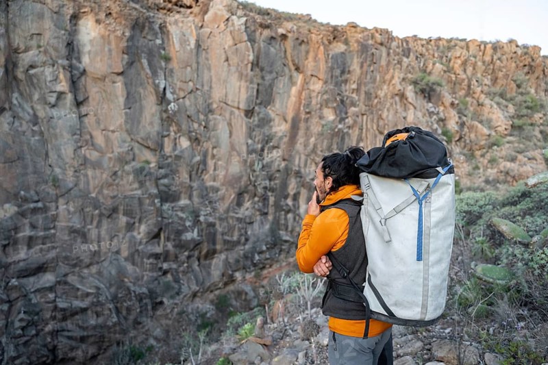 A climber with an orange jacket and a large backpack pauses to observe a towering rock face, contemplating the ascent. The scene captures the essence of traditional climbing
