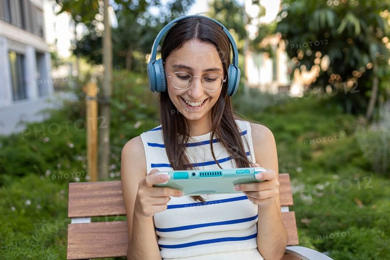 A young woman relaxes on a city park bench, wearing headphones, and playing a handheld game console. She embraces urban outdoor life, enjoying tranquility and technology