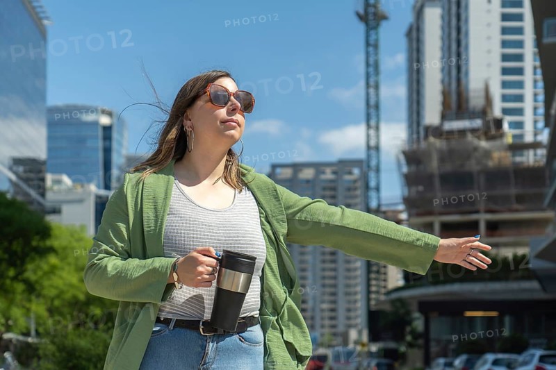 Businesswoman hailing a taxi in a modern city center, holding a coffee thermos and wearing sunglasses