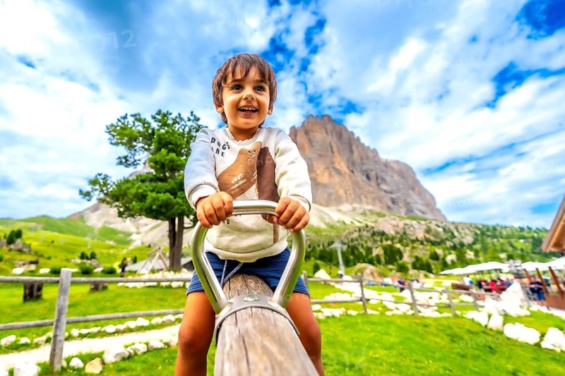 Happy child playing on a swing in a playground in the italian dolomites mountains, with the sassolungo peak in the background, during a sunny summer day