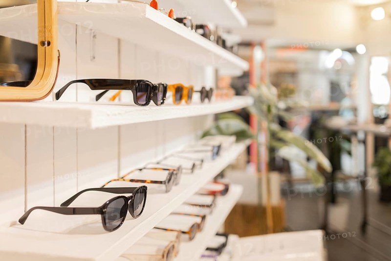 A stylish collection of sunglasses is displayed on white wooden shelves in a trendy retail shop, showcasing a variety of designs, colors, and frames suitable for all