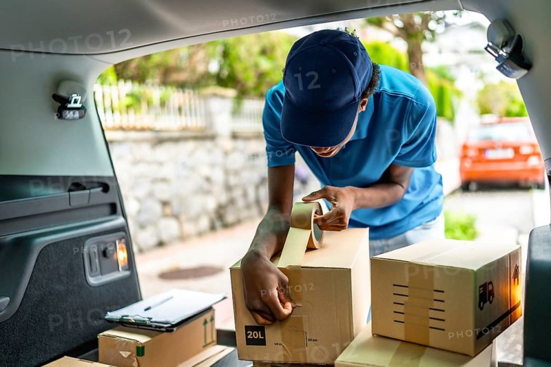 Delivery african young man sealing a cardboard package with tape in the van