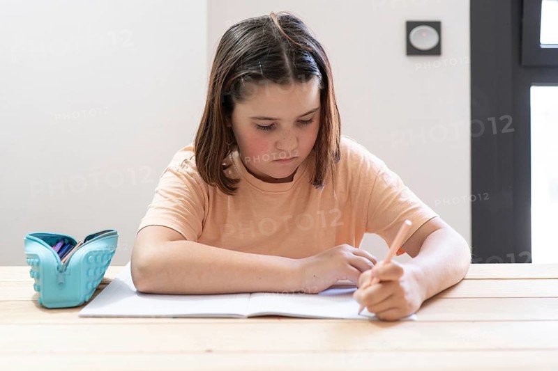 A young girl with brown hair concentrates on writing in her notebook. She sits at a wooden table with a blue pencil case, immersed in a quiet study session