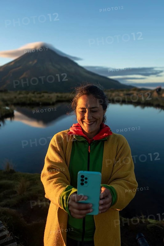 Smiling woman takes a selfie with a smartphone during sunrise at Mount Taranaki, Nueva Zelanda. The mountain's reflection creates a serene, picturesque scene