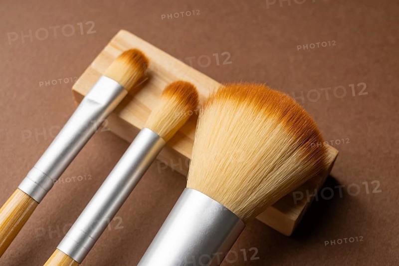 From above, three bamboo makeup brushes with silver metal accents rest on a light wood holder against a smooth brown background, showcasing eco-friendly design