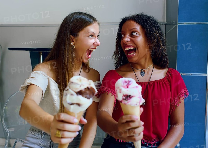 Two teenage girls, one Caucasian and one African descent, laughing joyfully while holding ice cream cones in a relaxed indoor setting