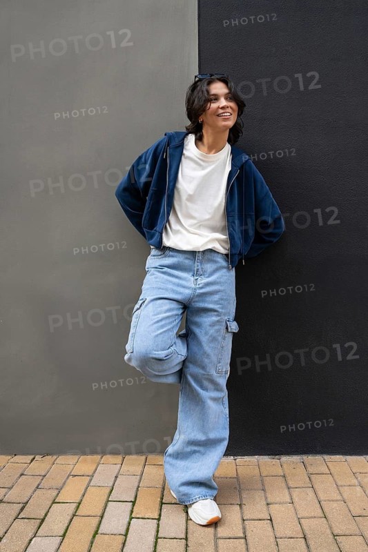 A person leans against a two-tone wall, wearing a casual outfit with a blue hoodie, white T-shirt, light blue jeans, and white sneakers. The scene exudes a relaxed urban vibe