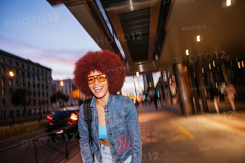 An African descendent woman with vibrant afro hair enjoys the evening, walking under city street lights. She wears stylish orange sunglasses and a denim jacket, her smile expressing pure joy
