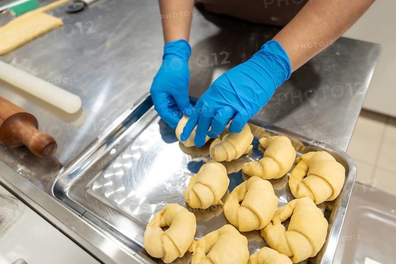 Professional baker hands wearing blue gloves preparing unbaked croissant and pastry dough on a metal tray, demonstrating food preparation and artisanal baking process in a commercial kitchen