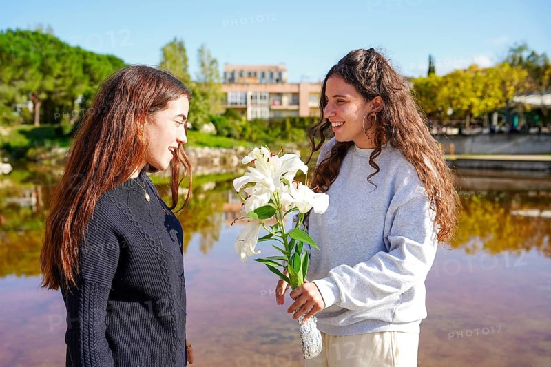 A romantic young happy women couple in the LGBTIQ+ community shares a moment by a serene pond, with one offering white lilies to the other amidst a sunny, natural setting