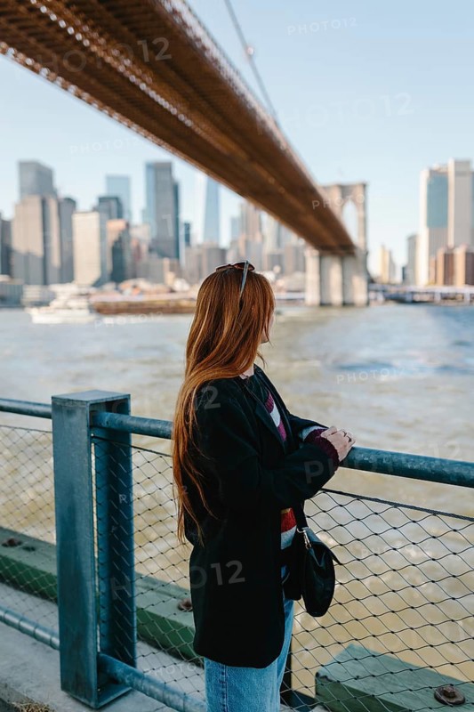 A woman with long hair gazes at the cityscape from beneath an iconic bridge in New York. Capturing urban life, the scene highlights architecture and serenity by the water