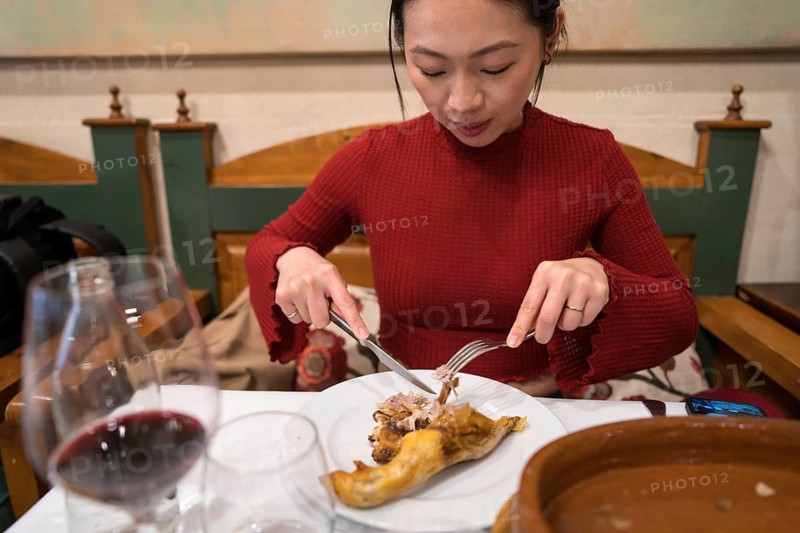 An Asian female tourist in Segovia, Spain, enjoys a traditional roast suckling pig dish. Seated in a cozy restaurant, she delicately samples the local culinary specialty