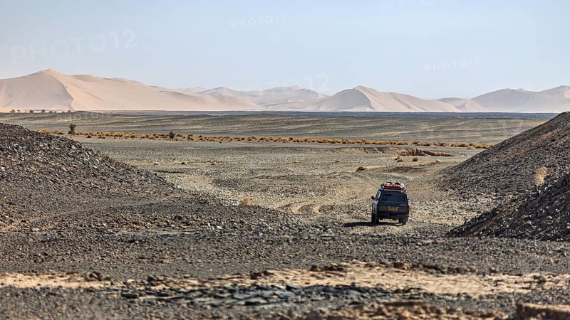 A vehicle moves through a barren desert landscape behind sand dunes, With off-road vehicle through the Sahara desert in Algeria