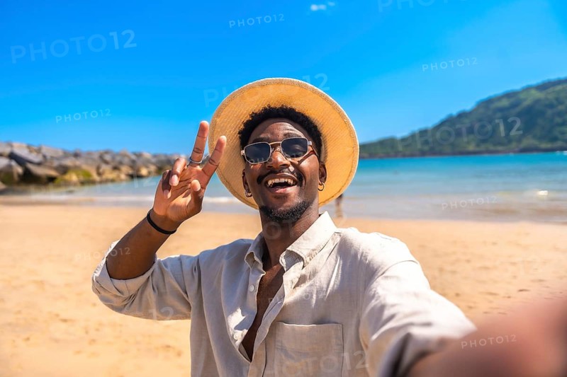 Young man wearing hat and sunglasses gesturing victory sign while taking selfie on a tropical beach