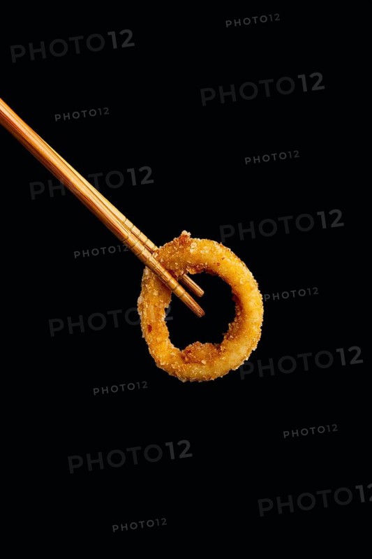 A golden, crispy onion ring delicately held between wooden chopsticks, set against a dark background. Highlights the texture and color contrast, ideal for culinary themes