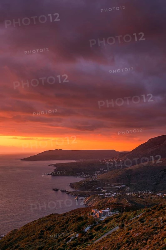 Dramatic sunset over the Mani Peninsula with vibrant orange and purple skies, highlighting coastal villages, rugged hills, and the shimmering sea along the Greek coastline