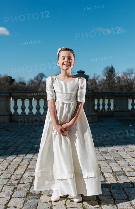 A young girl stands outdoors in a beautiful First Communion dress, radiating joy on a sunny day. The setting includes a clear blue sky and tree-lined background