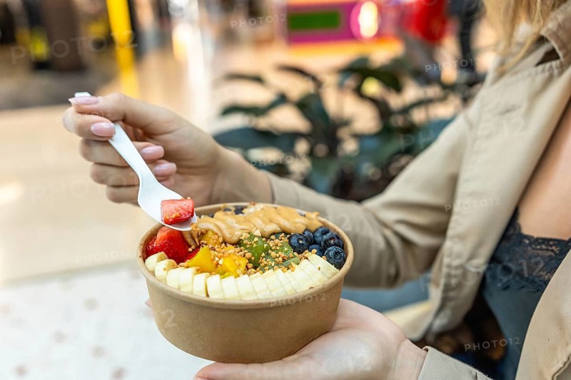 Woman holding and eating an acai bowl filled with fresh strawberries, bananas, blueberries, kiwi, and peanut butter, representing healthy eating and lifestyle