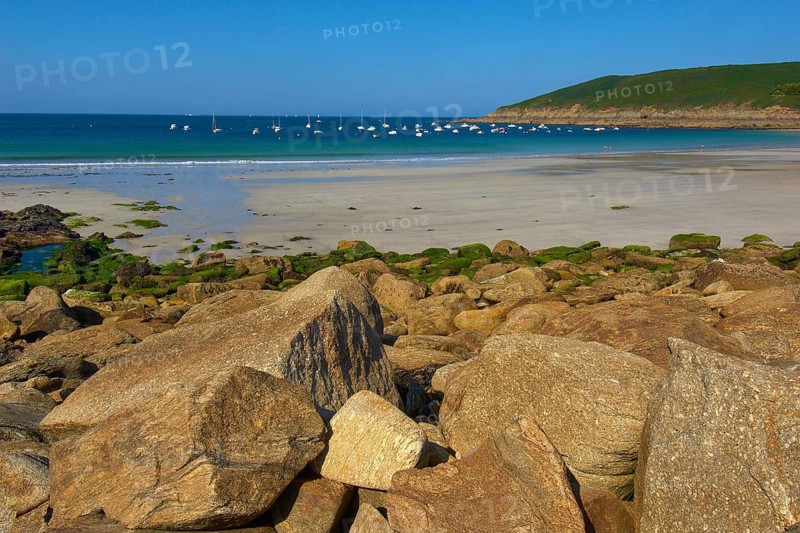 Quiet beach with many boats on the blue sea and rocks in the foreground, Brittany, France