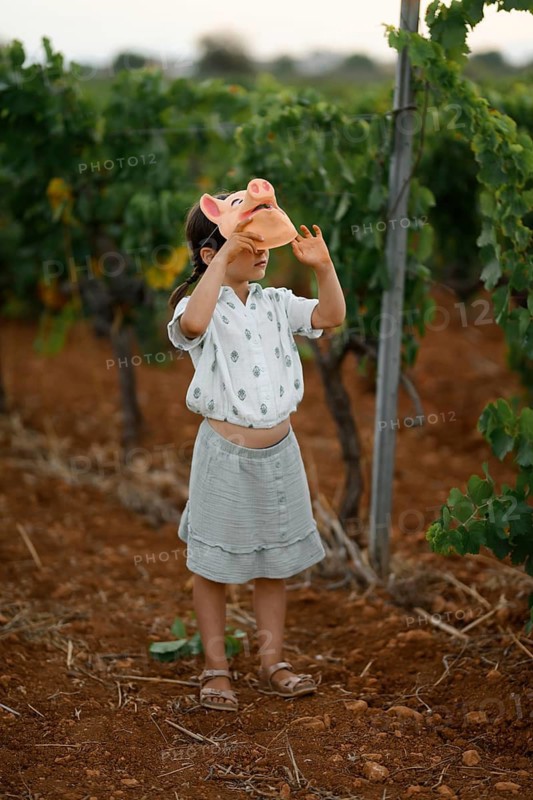 A young child stands in a vineyard, playfully holding a pig mask over their head. The scene highlights creativity and fun in a natural, sunny outdoor setting